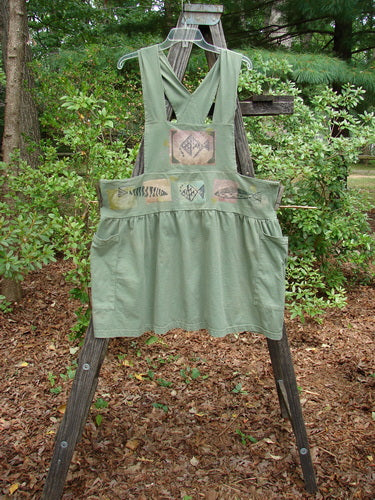 1995 Overall Jumper Fish Path Dinette Green OSFA displayed on a wooden ladder, featuring a creatively cut overall bib, crisscross back straps, and a full gathered skirt with oversized side pockets.