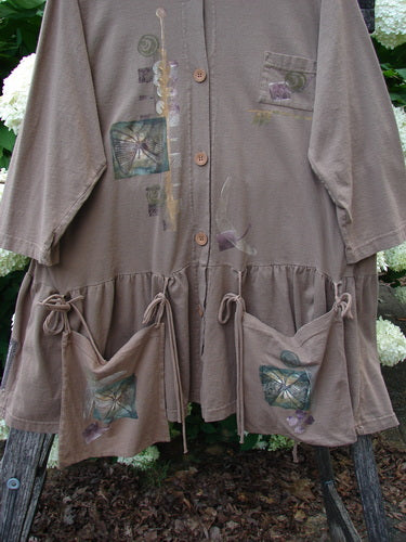 Bluefishfinder.com 1995 Clothespin Jacket Butterfly Mudpie OSFA in muted taupe, a long button-front jacket with a soft, draped knit texture. It features butterfly appliqués, a vertical floral motif, and a gathered hem with tied patch pockets, shown hanging outdoors among dark wood and pale flowers.