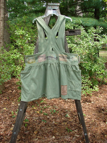 1995 Overall Jumper Fish Path Dinette Green OSFA displayed on a wooden pole, featuring a creatively cut overall bib, wide playful skirt, oversized side pockets, crisscross back straps, and fish-themed design.