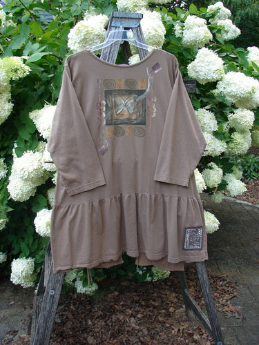 Bluefishfinder.com 1995 Clothespin Jacket Butterfly Mudpie OSFA in muted taupe, a loose long-sleeve cloth jacket with a butterfly graphic at the chest, gathered hem, and a small patch label near the lower right. It hangs from a weathered wooden stand against white hydrangeas and green foliage, with soft natural light.
