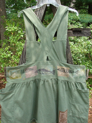 1995 Overall Jumper Fish Path Dinette Green OSFA: Green overalls with a fish design, featuring a creatively cut bib, wide skirt, crisscross back straps, and oversized side pockets, displayed on a wooden pole.