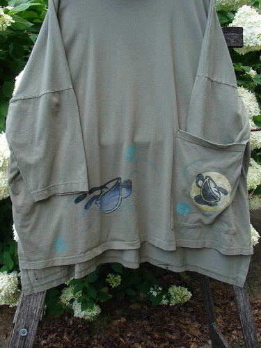 Bluefishfinder.com 1996 Message Top Tumbling Teacup Vine Size 3 in muted olive green, a loose long-sleeve top with oversized patch pockets and faded teacup vine graphics. The soft, worn fabric hangs in a relaxed drape against leafy greenery.