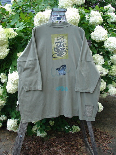 Bluefishfinder.com 1996 Message Top Tumbling Teacup Vine Size 3, an olive-gray long-sleeve shirt with a loose, boxy cut and dropped shoulders. A centered graphic panel, blue teacup motif, and small chest print sit on the soft cotton-like fabric, while the shirt hangs on a wooden stand among white hydrangeas.