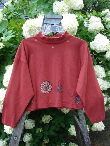 Bluefishfinder.com 1997 Structure Top Continuous Pinwheel Brick OSFA, a brick-red cropped long-sleeve top with a high mock neck and loose drape. Circular pinwheel motifs decorate the lower front and sleeves, while the soft fabric hangs from a wooden hanger amid white hydrangeas and green leaves.