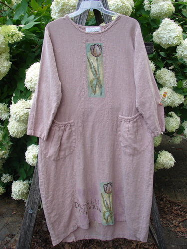 Bluefishfinder.com 1998 Botanicals Thostle Dress Single Tulip Mallow Size 1 in dusty mauve linen, a loose long-sleeve dress with a rounded neckline, side patch pockets, and vertical tulip-mallow appliqués on the front. It hangs on a clear hanger against white hydrangeas and green foliage.