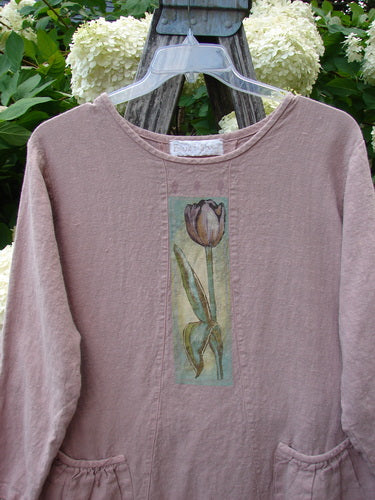 Bluefishfinder.com 1998 Botanicals Thostle Dress Single Tulip Mallow Size 1 in muted mauve, a long-sleeve dress with a round neckline, gathered waist, and front patch pockets. A narrow botanical panel with a tulip-mallow illustration runs down the chest, and the soft, textured fabric hangs from a clear hanger among pale hydrangea blooms and weathered wood.