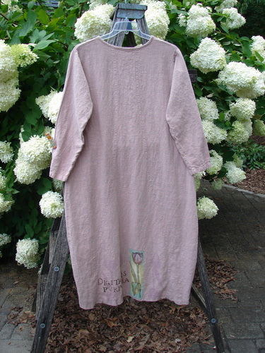Bluefishfinder.com 1998 Botanicals Thostle Dress Single Tulip Mallow Size 1 in muted mauve linen, a loose long-sleeve dress with a round neckline and a small floral print near the hem. It hangs centered on a wooden frame before white hydrangeas and dense green foliage.