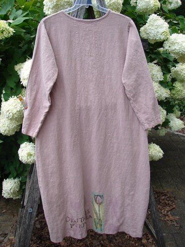 Bluefishfinder.com 1998 Botanicals Thostle Dress Single Tulip Mallow Size 1 in muted mauve linen, hanging front-facing with long sleeves, a simple round neckline, and a soft, textured drape. A small botanical print near the hem adds a delicate vertical accent against the pale, natural fabric.