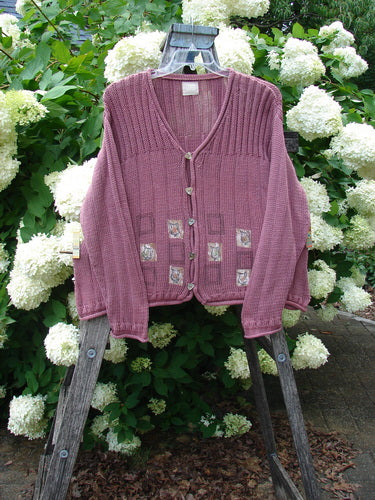 Bluefishfinder.com 1999 Triangle Cardigan Sweater Pitcher Clay Size 1 in muted pink-purple knit, with a deep V-neck, ribbed trim, and small patterned square patches near the hem. It hangs on a weathered wooden ladder before dense white hydrangeas and green foliage.