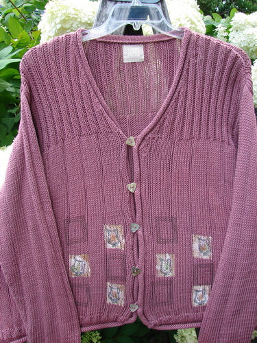 Bluefishfinder.com 1999 Triangle Cardigan Sweater Pitcher Clay Size 1 in muted mauve knit, with a deep V-neck, ribbed texture, and small triangular buttons down the front. Five square patch details sit on the lower panels, while the sweater hangs on a clear hanger against soft white flowers and green foliage.