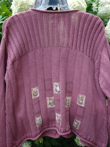 Bluefishfinder.com 1999 Triangle Cardigan Sweater Pitcher Clay Size 1 in a muted mauve-purple knit, with a ribbed yoke, relaxed long sleeves, and a softly gathered hem. Small patterned patches decorate the lower front, while the sweater hangs on a hanger against leafy greenery and white flowers.