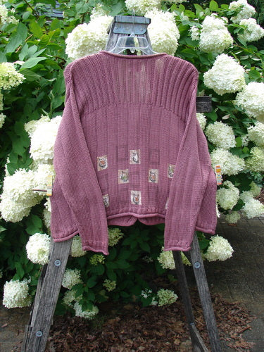Bluefishfinder.com 1999 Triangle Cardigan Sweater Pitcher Clay Size 1 in dusty mauve, a ribbed knit cardigan with a wide yoke, long sleeves, and small patchwork triangle motifs near the hem. It hangs from a wooden ladder against dense white hydrangeas and green foliage, with soft daylight and earthy tones.