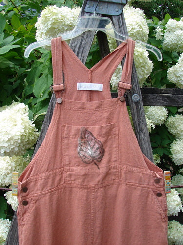 Bluefishfinder.com 1998 Botanicals Planters Overall Jumper Leaf Mimosa Size 0 in dusty rose fabric, with a V-neck bib, adjustable shoulder straps, and a front leaf-print pocket. It hangs from a clear hanger against weathered wood and white hydrangeas.