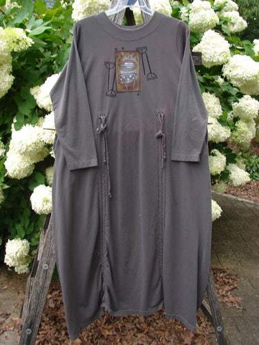 Bluefishfinder.com 1998 Cornucopia Dress Fall Garden Eggplant Size 1 in a muted eggplant tone, with a loose long-sleeve silhouette, gathered side ties, and a small centered graphic at the chest. The soft, draping fabric hangs from a wooden stand, set against dense white hydrangeas and green foliage.