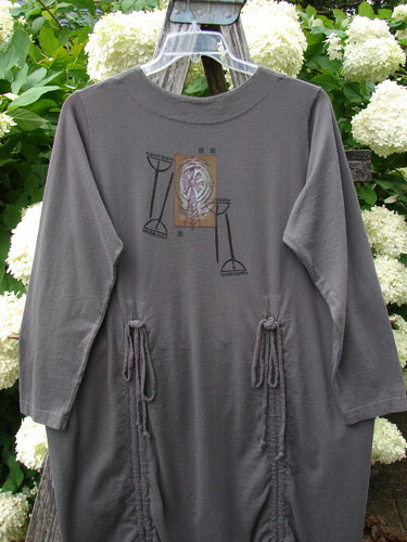 Bluefishfinder.com 1998 Cornucopia Dress Fall Garden Eggplant Size 1 in muted eggplant gray, a long-sleeve dress with a round neckline, central graphic print, and gathered side ties. It hangs on a clear hanger against white hydrangeas and weathered wood.