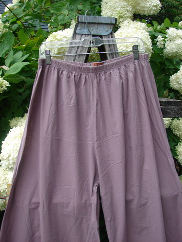 Bluefishfinder.com Barclay Batiste Tiny Flutter Pocketless Pant in unpainted dusty plum, shown as loose, softly draped pants with a gathered elastic waistband and a smooth, pocketless front. The matte fabric hangs from a hanger against leafy white hydrangeas, giving the image a natural, muted palette.