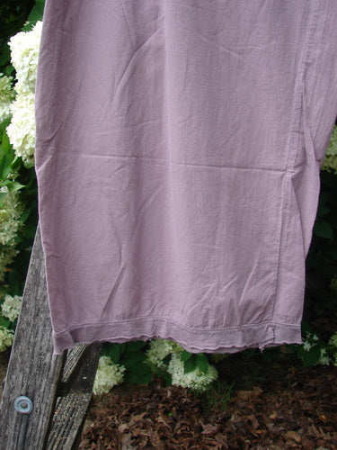 Bluefishfinder.com Barclay Batiste Tiny Flutter Pocketless Pant in unpainted dusty plum, shown as a loose, softly wrinkled pant leg with a fluttered hem and pocketless front. The lightweight fabric hangs against leafy greenery and white flowers, with muted light emphasizing its washed texture.