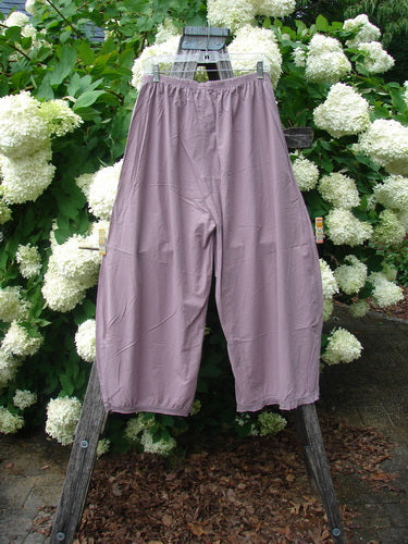 Bluefishfinder.com Barclay Batiste Tiny Flutter Pocketless Pant in unpainted dusty plum, a loose, cropped pair of pants with a gathered elastic waist and soft fluttered hems. The lightweight fabric hangs from a wooden stand, set against dense white blossoms and green foliage.