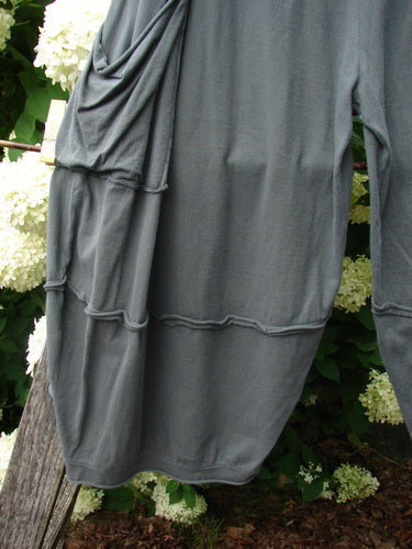 Bluefishfinder.com Barclay Crop 2 Pocket Barrel Pant in Unpainted Steel Night, shown in a soft charcoal-gray fabric with a loose barrel shape, cropped hem, and visible side pocket seams. The draped garment hangs against leafy white flowers and weathered wood.