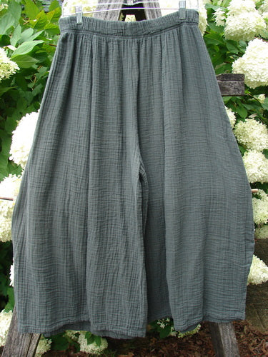 Bluefishfinder.com Barclay Gauze Crop Mojo Pant, unpainted evergreen size 1, shown as loose, cropped gauze pants with a softly crinkled texture, elastic waistband, and wide drape. The muted green fabric hangs against leafy white blossoms, emphasizing its airy, matte finish.