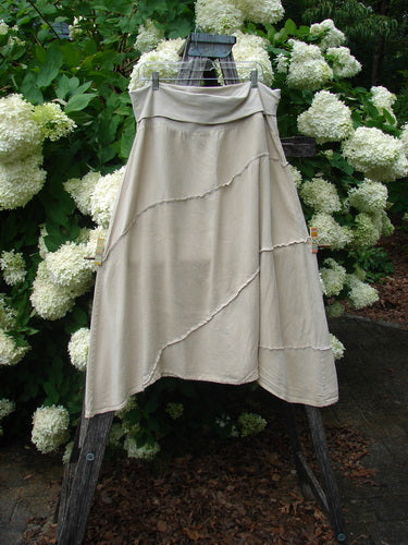 Barclay Hemp Silk Fold Over Diagonal Skirt in unpainted wheat, size 2, with a soft draped fold-over waistband and layered diagonal seams. The light hemp-silk fabric hangs in a fluid A-line shape against lush white hydrangeas and dark wood.