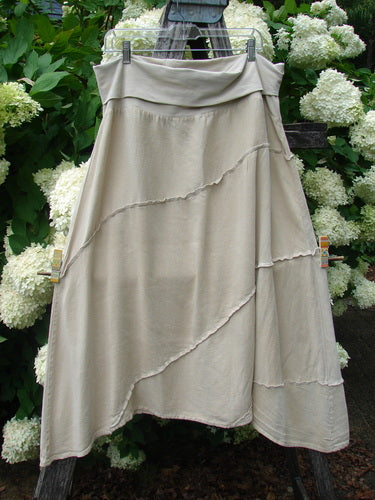 Barclay Hemp Silk Fold Over Diagonal Skirt in unpainted wheat, size 2, with a soft draped fold-over waistband and layered diagonal seams. The lightweight hemp-silk fabric hangs in a pale beige silhouette against dense white hydrangeas and green foliage.