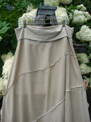 Barclay Hemp Silk Fold Over Diagonal Skirt in unpainted wheat, size 2, with a soft draped fold-over waistband and diagonal raw-edged seams. The lightweight hemp-silk fabric hangs in a flowing A-line shape, shown on a hanger against white flowers and dark wood.