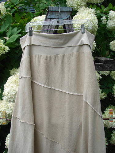 Barclay Hemp Silk Fold Over Diagonal Skirt in unpainted wheat, size 2, with a soft draped fold-over waistband and layered diagonal panels. The hemp-silk fabric has a matte, natural texture in pale beige, hanging from a clear hanger against dense green foliage and white blooms.