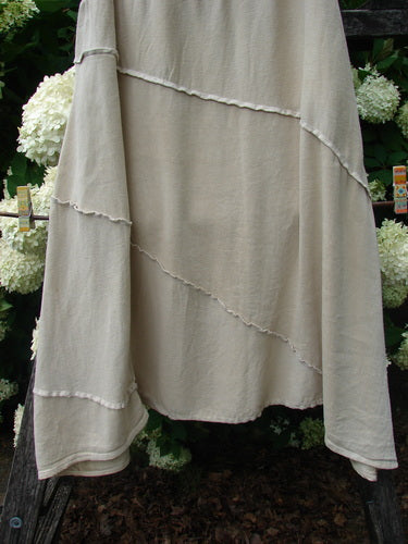 Barclay Hemp Silk Fold Over Diagonal Skirt in unpainted wheat, size 2, with layered diagonal panels and raw-edged seams in a soft hemp-silk drape. The long skirt hangs in a muted beige tone against dark greenery and white flowers.