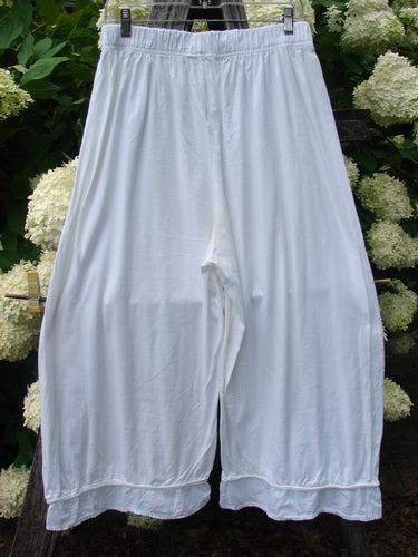 Barclay Flutter Bottom Pocketless Pant in unpainted white, size 1, with a loose, wide-leg silhouette and soft fluttered hems. The lightweight fabric hangs in smooth folds, centered against dark wood and pale hydrangea blooms.