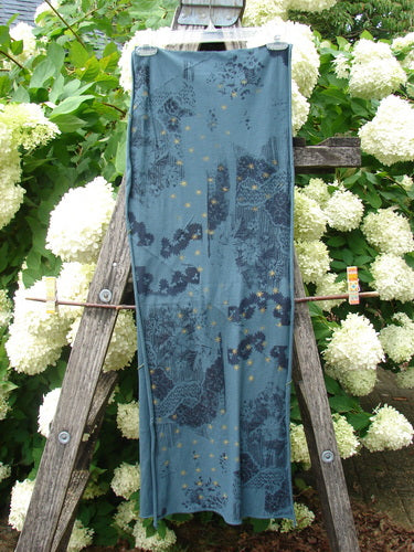Bluefishfinder.com Barclay Diagional Scarf Wrap Stary Night Garden Blue Hunter OSFA is a long, sheer blue wrap with a diagonal floral-and-nightscape print in darker navy tones and scattered yellow specks. It hangs vertically over a weathered wooden ladder, its lightweight fabric falling in soft folds against dense white blossoms and green foliage.