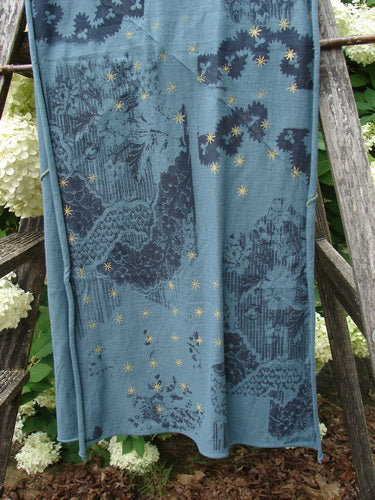 Bluefishfinder.com Barclay Diagional Scarf Wrap Stary Night Garden Blue Hunter OSFA drapes in a blue-green field, patterned with dark botanical panels and scattered gold star motifs. The lightweight fabric hangs in soft vertical folds, creating a layered, textured look against the rustic wooden backdrop.