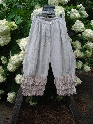 Magnolia Pearl Homespun Linen Ruffle Eyelet Bloomer in natural, an OSFA pair of loose, ankle-length bloomers with a soft linen texture, gathered waist, and layered ruffle eyelet hems. They hang centered on a wooden ladder amid white hydrangeas and deep green foliage.