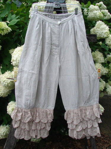 Magnolia Pearl Homespun Linen Ruffle Eyelet Bloomer in natural, shown as airy linen bloomers with a softly gathered waist, button front, and layered eyelet ruffles at the cropped hems. The pale neutral fabric hangs with a loose, textured drape against dense green foliage and white hydrangeas.