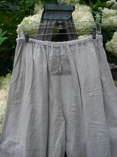 Magnolia Pearl Homespun Linen Ruffle Eyelet Bloomer in natural OSFA, shown in soft gray-beige linen with a gathered elastic waistband and loose, airy drape. A faded front patch sits near the center, while the garment hangs from a clear hanger against dark wood and pale hydrangeas.