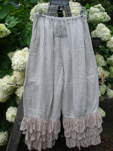 Magnolia Pearl Homespun Linen Ruffle Eyelet Bloomer in natural OSFA, shown as loose, pale linen bloomers with an elastic waist and layered eyelet ruffles at the hems. The soft, rumpled fabric hangs from a hanger against dense white hydrangeas and dark greenery.