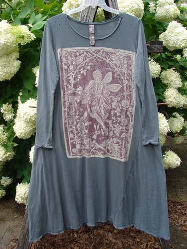 Bluefishfinder.com Magnolia Pearl Cotton Jersey Dylan Dress Fairy Ozzy OSFA in faded blue cotton jersey, with a loose long-sleeve silhouette, raw edges, and a large pink floral graphic panel on the front. It hangs centered against lush white hydrangeas and green foliage, giving the garment a soft, rustic outdoor presentation.