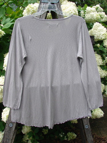 Bluefishfinder.com Barclay Modal Slub Long Sleeved A Lined Tee in peacock grey, size 0, shown front-on with a softly draped A-line silhouette, long sleeves, and a gently rounded neckline. The modal slub fabric has a fine textured finish, and the hem and cuffs are left with a slightly raw, wavy edge against a leafy outdoor backdrop.