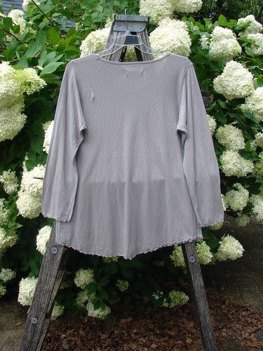 Bluefishfinder.com Barclay Modal Slub Long Sleeved A Lined Tee in peacock grey, size 0, hangs from a weathered wooden easel. The soft modal slub knit has a relaxed A-line shape, long sleeves, and a gently scalloped hem, set against dense white hydrangeas and green foliage.