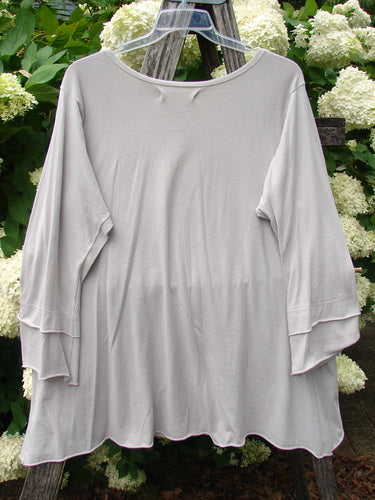 Bluefishfinder.com Barclay Petal Sleeve A Line Tunic Falling Fern Birch Bark Size 1 in birch bark, a softly draped light gray tunic with petal sleeves, a rounded neckline, and an A-line silhouette. It hangs from a clear hanger against white hydrangeas and weathered wood.