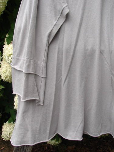 Bluefishfinder.com Barclay Petal Sleeve A Line Tunic in Falling Fern Birch Bark, a soft birch-bark gray knit with layered petal sleeves and a gently flared A-line hem. The fabric drapes loosely in the foreground, with dark foliage and pale flowers behind it.