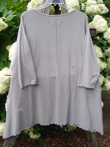 Barclay Cotton Lycra 2 Pocket Urchin Dress Flower Tops Size 0 in soft gray cotton Lycra, a loose long-sleeve dress with a round neckline, raw scalloped hems, and a straight drape. It hangs centered against leafy white flowers, emphasizing its lightweight, matte texture.