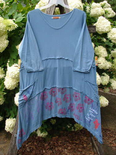 Bluefishfinder.com Barclay Raw Seam Vector Tunic Dress Daisy Folly Echo Blue Size 1, a loose blue tunic dress with raw seam layers, long sleeves, and an asymmetric hem. The lower panel shows a darker floral print with white graphic text, photographed hanging outdoors against leafy white blossoms.