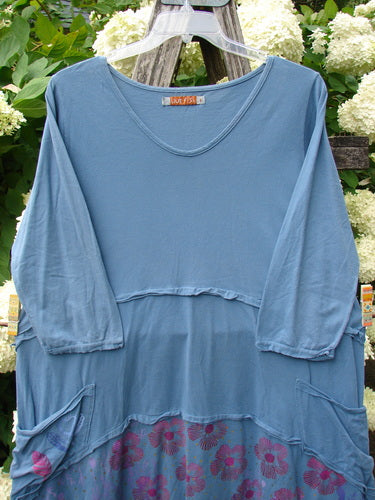 Bluefishfinder.com Barclay Raw Seam Vector Tunic Dress Daisy Folly Echo Blue, a loose blue tunic dress with raw seam detailing, three-quarter sleeves, and a softly gathered waist. The lower hem reveals a daisy folly floral print, while the garment hangs on a hanger against leafy white blossoms.
