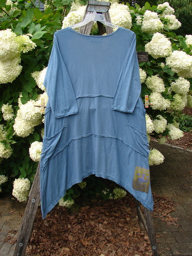Bluefishfinder.com Barclay Raw Seam Vector Tunic Dress Daisy Folly Echo Blue Size 1 is a loose, draped blue tunic dress with raw seams, a rounded neckline, and layered curved panels. The soft, washed fabric hangs from a wooden stand, with white hydrangeas framing it behind.