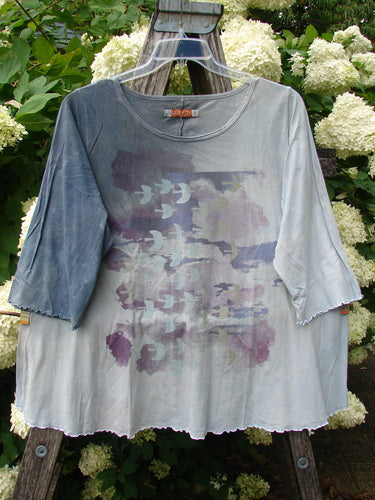 Bluefishfinder.com Barclay Three Quarter Sleeved Cafe Tee in Dove Flight mottled grey blue cloud, a loose cropped top with raglan three-quarter sleeves and a raw, uneven hem. Its soft, lightweight fabric shows a faded bird print in muted lavender, blue, and grey tones.