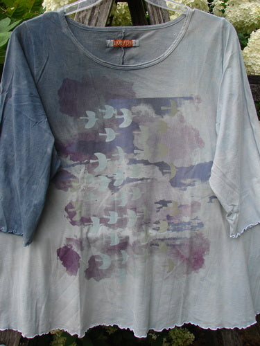 Bluefishfinder.com Barclay Three Quarter Sleeved Cafe Tee in Dove Flight Mottled Grey Blue Cloud, a loose crewneck top with three-quarter sleeves and a soft, washed finish. The front shows layered dove graphics in muted purple, gray, and blue tones.