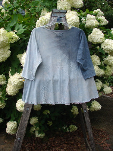 Bluefishfinder.com Barclay Three Quarter Sleeved Cafe Tee in Dove Flight mottled grey blue cloud, a soft, lightweight top with three-quarter sleeves, a gently flared hem, and a raw-edged finish. It hangs from a wooden ladder before creamy white hydrangeas, the cool blue-gray fabric contrasting with the lush green foliage.