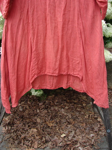 Bluefishfinder.com Barclay Linen High Vent Venetian Dress Center Starburst Poppy Size 1 in a coral-poppy linen drape, with a high center vent and softly crinkled texture. The fabric falls in a loose, layered silhouette over a dark frame, with mulch and pale flowers visible below.
