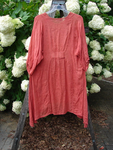 Bluefishfinder.com Barclay Linen High Vent Venetian Dress Center Starburst Poppy Size 1, a coral-poppy linen dress with a soft crinkled texture, loose long sleeves, and a gently gathered center seam. It hangs on a wooden stand before white hydrangeas, with muted natural light and earthy ground tones.